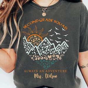 May include: A charcoal grey t-shirt featuring a graphic print with the text "SECOND GRADE SQUAD" and "ALWAYS AN ADVENTURE Mrs. Wilson". The design includes a sun, mountain peaks filled with floral patterns, and a flock of birds in flight.