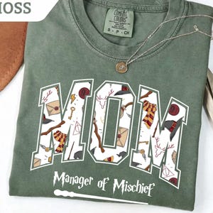 May include: A moss green t-shirt featuring the word MOM in large block letters filled with a pattern of wizarding-themed icons including wands, scarves, and letters. Below the word, the text reads Manager of Mischief with a wand graphic.