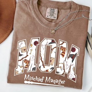 May include: Espresso-colored t-shirt with the word "MOM" in a white, outlined font, filled with themed illustrations. Below, the text "Mischief Manager" is displayed. A silver necklace with a pendant is visible.