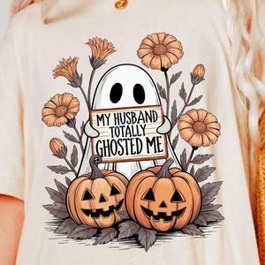 May include: Ivory-colored t-shirt featuring a Halloween-themed graphic. The design includes a white ghost holding a sign that reads "MY HUSBAND TOTALLY GHOSTED ME." Two carved pumpkins and orange flowers surround the ghost.