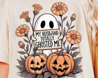 May include: Ivory-colored t-shirt featuring a Halloween-themed graphic. The design includes a white ghost holding a sign that reads "MY HUSBAND TOTALLY GHOSTED ME." Two carved pumpkins and orange flowers surround the ghost.