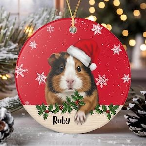 Könnte beinhalten: Ein runder Weihnachtsbaumschmuck aus Keramik mit einem braun-schwarz-weißen Meerschweinchen, das eine rot-weiße Weihnachtsmütze trägt. Der Hintergrund ist rot mit weißen Schneeflocken, unten sind grüne Stechpalmenblätter und der Name Ruby in Schwarz.