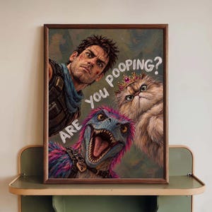 May include: A framed wall art print featuring a man with dark hair, a fluffy cat wearing a gold crown, and a blue and pink feathered dinosaur. The text "Are you pooping?" is written in white script across the centre of the colourful illustration.