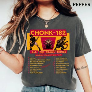 May include: A charcoal gray t-shirt featuring a graphic print with the text CHONK-182 ALL THE CRAWL THINGS DUNGEON CRAWLER WORLD TOUR. The design includes a guitarist, a drummer, and a dinosaur playing guitar, with a list of tour locations below.