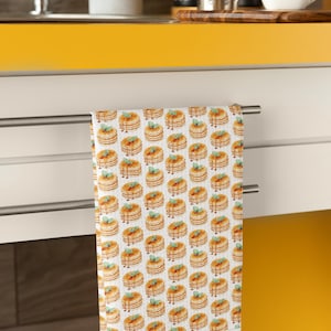 May include: A white kitchen towel with a repeating pattern of stacks of pancakes with syrup and a sprig of mint on top.