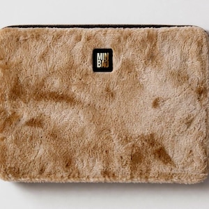 May include: A rectangular, tan-colored laptop sleeve with a plush, furry texture. The sleeve has a black zipper along the top edge and a small, square black label with gold lettering in the center.