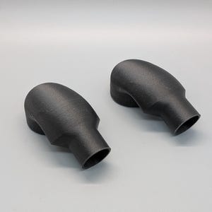 May include: Two black, rounded, 3D-printed objects with a textured surface. Each object has a bulbous top and a cylindrical base. The objects are positioned on a light gray surface.