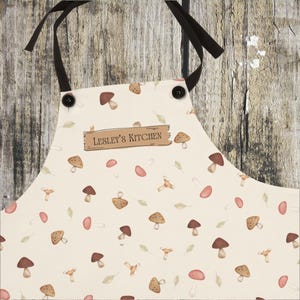 May include: A white apron with a mushroom pattern and the text "Lesley's Kitchen" on a brown label. The apron has black straps and two black buttons.