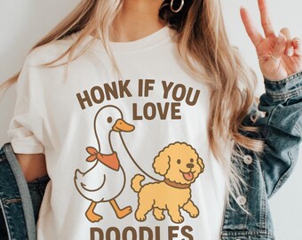 May include: White t-shirt with a cartoon duck wearing an orange bandana walking a yellow doodle dog on a leash. The text "HONK IF YOU LOVE DOODLES" is printed above and below the illustration in brown.