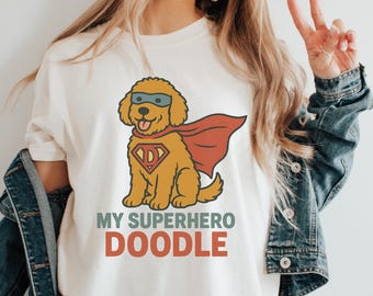 May include: Cream-colored t-shirt featuring a cartoon doodle dog dressed as a superhero, complete with a cape and mask. The text "MY SUPERHERO DOODLE" is printed below the dog in green and red. The dog has a shield with the letter "D".
