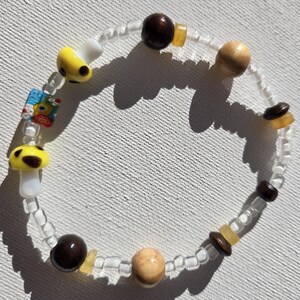 May include: A beaded bracelet featuring a variety of beads. The bracelet includes mushroom-shaped beads with yellow caps and brown spots, wooden beads, and small clear beads. There are also small, square, colorful glass beads.