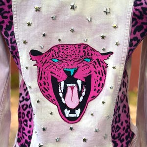 May include: Pink denim jacket with a large, roaring pink jaguar face on the back, accented with blue eyes and a black mouth. The jacket features leopard print panels and silver star-shaped studs.