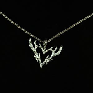 May include: A silver pendant necklace with a heart-shaped, textured design resembling branches. The pendant hangs from a fine silver chain. The necklace is set against a black backdrop. A piece of jewellery.