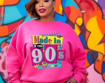 Puede incluir: Una sudadera rosa brillante con el texto "Made in the 90s" en un diseño retro colorido. La sudadera se combina con una gorra de béisbol roja y pantalones negros. El fondo presenta un vibrante mural de graffiti.