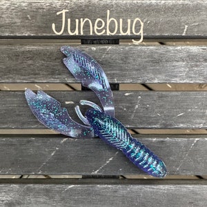 May include: A blue and green plastic crawfish lure with a glittery finish. The lure is designed to resemble a crawfish and is used for fishing.