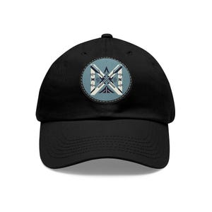 Op de afbeelding: Zwarte baseballpet met een gebogen klep. De pet heeft een cirkelvormige patch met een ontwerp van gekruiste roeispanen in wit en lichtblauw, tegen een lichtblauwe achtergrond. De letters "M" en "N" zijn zichtbaar op de patch.