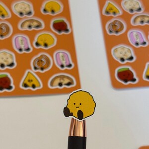 May include: A sticker sheet with an orange background featuring various small cartoon food illustrations with faces. A single yellow, round, textured food sticker with a smiling face and dark brown limbs is held up in the foreground on a pen tip.