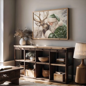 May include: A framed print depicts Santa Claus embracing a reindeer in a snowy scene. The artwork is in a wooden frame and hangs above a wooden console table with drawers and open shelves. The room is decorated with a lamp, vases, and baskets.