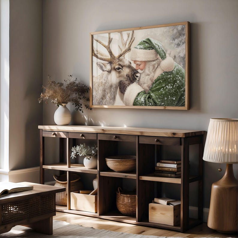 May include: A framed print depicts Santa Claus embracing a reindeer in a snowy scene. The artwork is in a wooden frame and hangs above a wooden console table with drawers and open shelves. The room is decorated with a lamp, vases, and baskets.