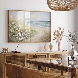 May include: A framed seascape painting with a beach scene and white flowers in the foreground. The painting is hanging on a white wall above a wooden cabinet with woven rattan doors.