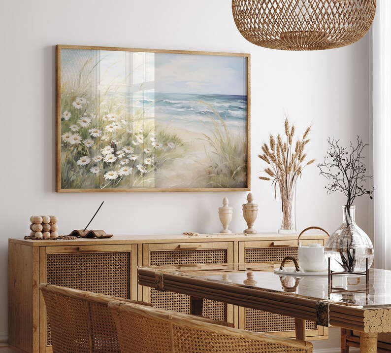 May include: A framed seascape painting with a beach scene and white flowers in the foreground. The painting is hanging on a white wall above a wooden cabinet with woven rattan doors.