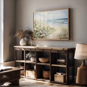 May include: A wooden shelf unit with six shelves, featuring a painting of a beach scene with white flowers, a vase with dried flowers, a bowl, a basket, and a book stack. The shelf unit is against a light gray wall.