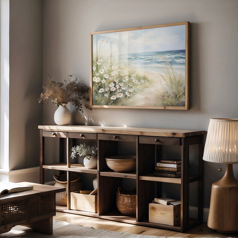 May include: A wooden shelf unit with six shelves, featuring a painting of a beach scene with white flowers, a vase with dried flowers, a bowl, a basket, and a book stack. The shelf unit is against a light gray wall.