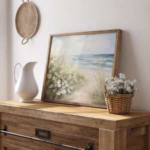 May include: A framed watercolor painting of a beach scene with white flowers and blue waves. The painting is on a wooden dresser with a white pitcher and a basket of white flowers.