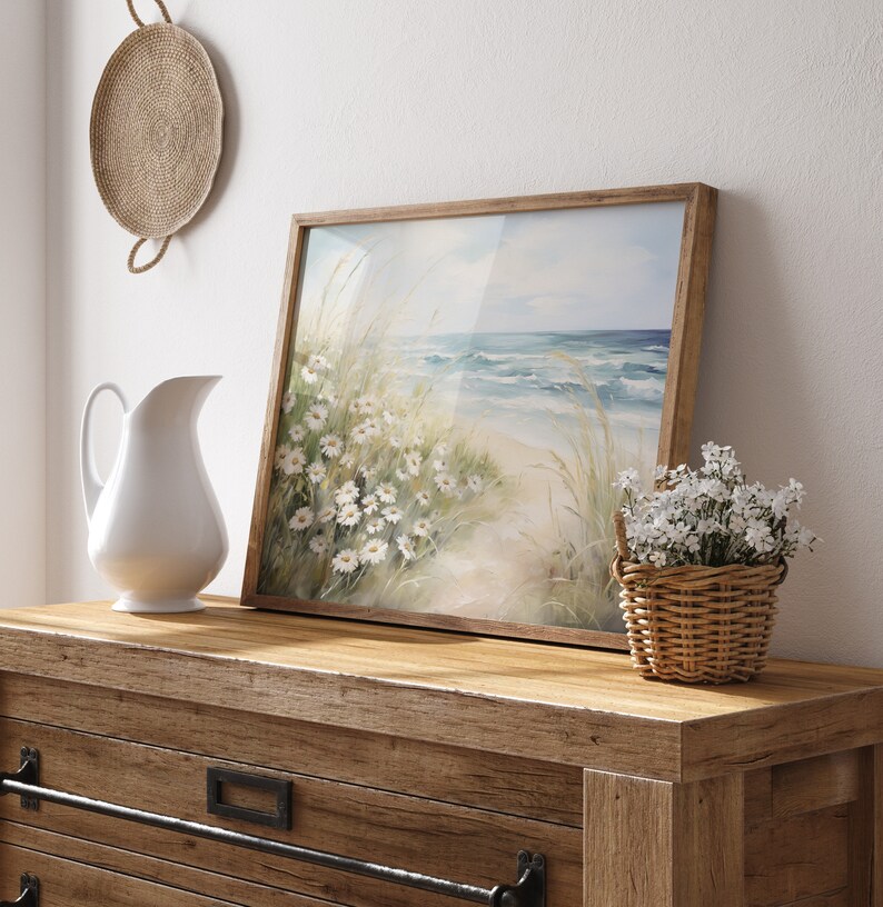 May include: A framed watercolor painting of a beach scene with white flowers and blue waves. The painting is on a wooden dresser with a white pitcher and a basket of white flowers.