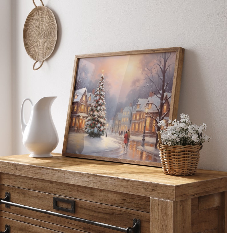 Puede incluir: Una pintura enmarcada de una escena de invierno con un pueblo nevado y un &aacute;rbol de Navidad iluminado en el centro. La pintura est&aacute; sobre una c&oacute;moda de madera con una jarra blanca y una cesta de mimbre con flores blancas.