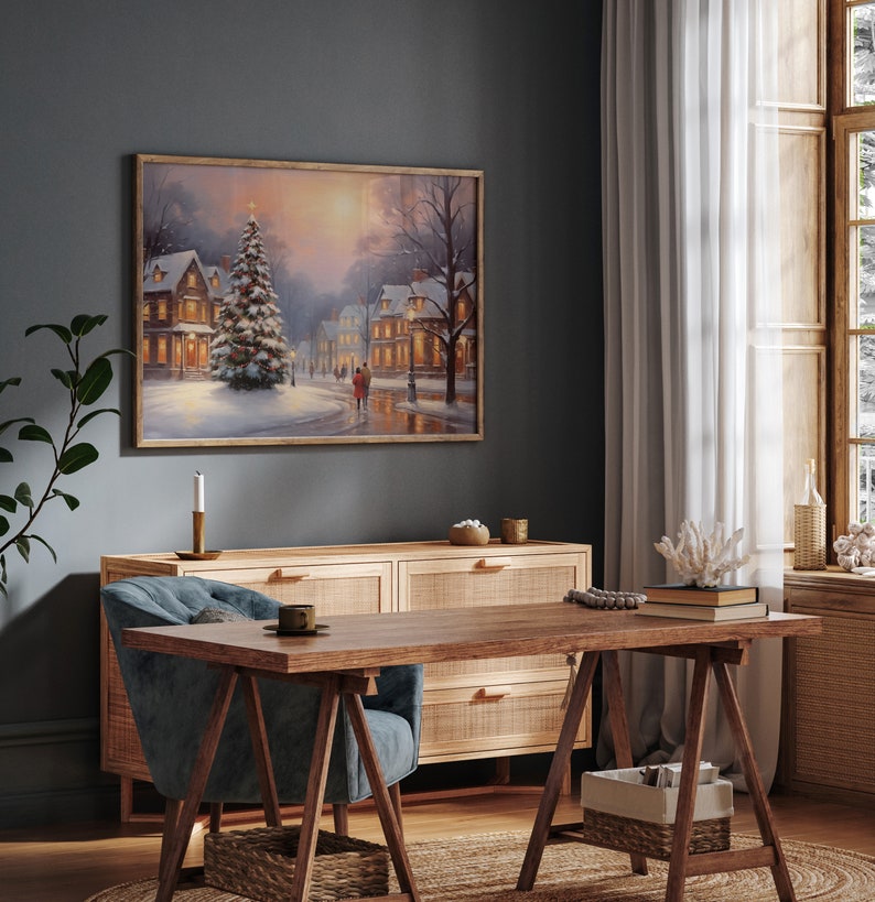 Puede incluir: Una pintura enmarcada que representa una escena de invierno nevada con un &aacute;rbol de Navidad iluminado en el centro, rodeado de casas con ventanas iluminadas. La pintura est&aacute; colgada en una pared gris oscura sobre una c&oacute;moda de madera con cajones.