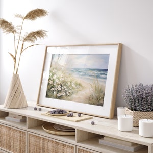 May include: A framed watercolor painting of a beach scene with white flowers and blue waves. The painting is on a wooden shelf with a vase of dried reeds, candles, and a bowl of blueberries.