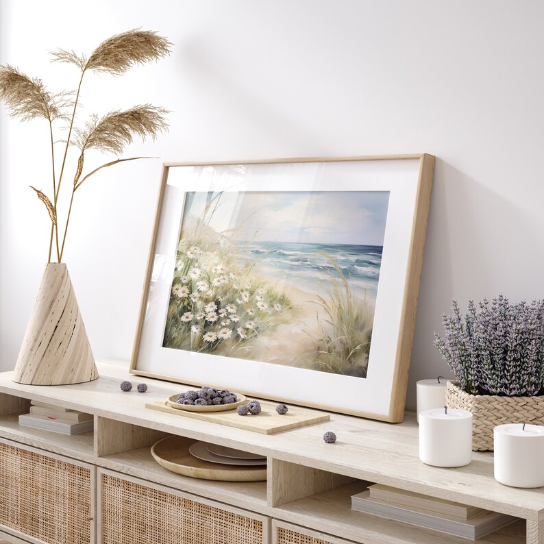 May include: A framed watercolor painting of a beach scene with white flowers and blue waves. The painting is on a wooden shelf with a vase of dried reeds, candles, and a bowl of blueberries.