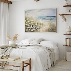 May include: A white bed with a white and gray blanket and a white pillow. A painting of a beach scene with white flowers hangs above the bed.
