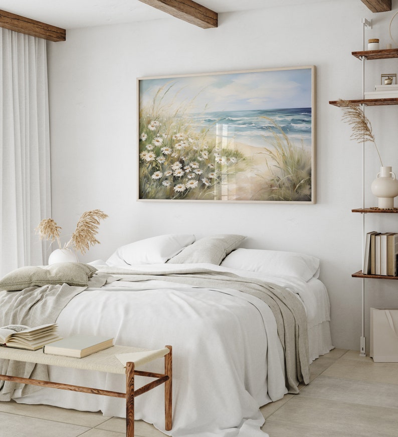 May include: A white bed with a white and gray blanket and a white pillow. A painting of a beach scene with white flowers hangs above the bed.