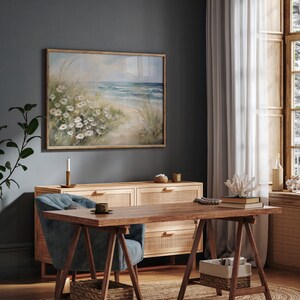 May include: A light blue armchair with a wooden frame sits in front of a wooden dresser with woven cane drawers. A framed watercolor painting of a beach scene with white flowers hangs on the wall behind the dresser. A wooden table with a rustic trestle base sits in front of the armchair.