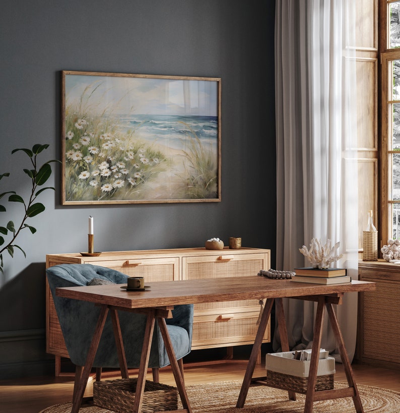 May include: A light blue armchair with a wooden frame sits in front of a wooden dresser with woven cane drawers. A framed watercolor painting of a beach scene with white flowers hangs on the wall behind the dresser. A wooden table with a rustic trestle base sits in front of the armchair.