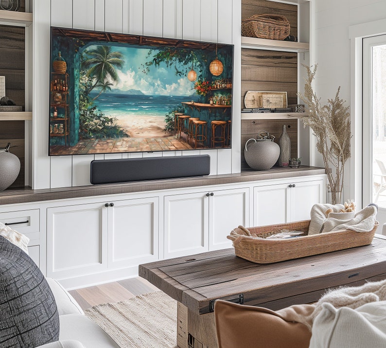 May include: A living room with a white and wood entertainment unit featuring a flat screen TV displaying a tropical beach scene. The entertainment unit has a soundbar and white cabinets. A wooden coffee table with a woven basket and a throw blanket sits in front of the entertainment unit.