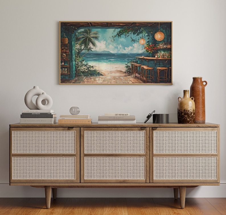 May include: A framed painting depicting a tropical bar scene with palm trees, a beach, and turquoise water. The bar has a thatched roof and is decorated with fairy lights. The painting is hanging on a white wall above a wooden cabinet with woven cane doors.