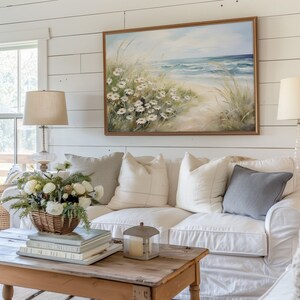 May include: A white couch with a variety of pillows, including a gray pillow, sits in front of a white shiplap wall. A framed painting of a beach scene with daisies hangs above the couch. A wooden coffee table with a stack of books and a candle lantern sits in front of the couch.