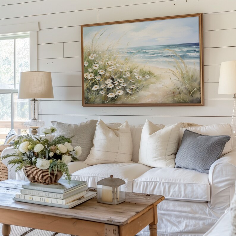 May include: A white couch with a variety of pillows, including a gray pillow, sits in front of a white shiplap wall. A framed painting of a beach scene with daisies hangs above the couch. A wooden coffee table with a stack of books and a candle lantern sits in front of the couch.