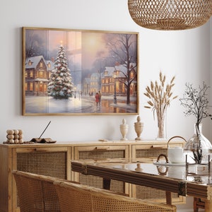Puede incluir: Una pintura enmarcada que representa una escena de invierno con un pueblo cubierto de nieve y un &aacute;rbol de Navidad en primer plano. La pintura est&aacute; colgada en una pared blanca sobre un armario de madera con puertas de mimbre.