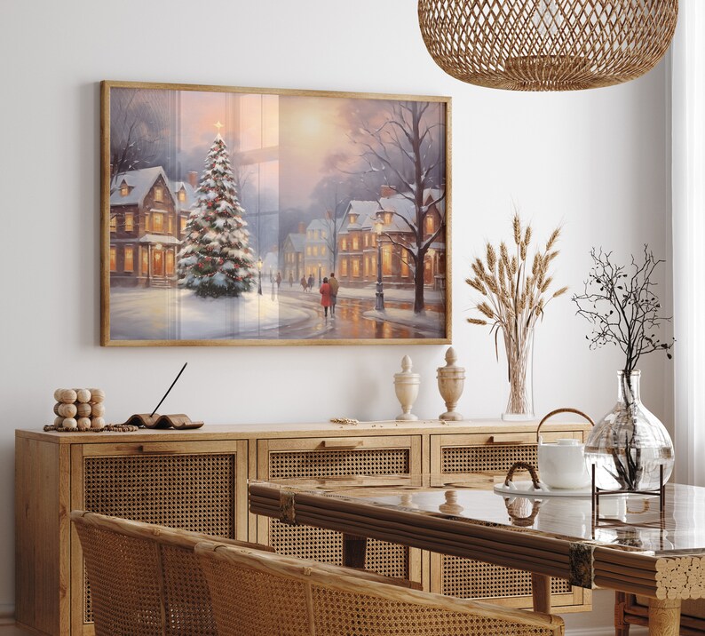 Puede incluir: Una pintura enmarcada que representa una escena de invierno con un pueblo cubierto de nieve y un &aacute;rbol de Navidad en primer plano. La pintura est&aacute; colgada en una pared blanca sobre un armario de madera con puertas de mimbre.