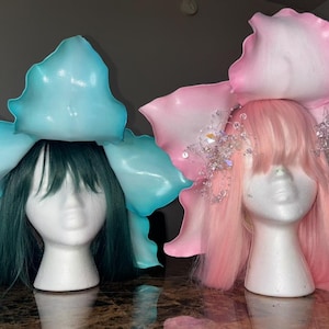 K&ouml;nnte beinhalten: Zwei Mannequin-K&ouml;pfe, die gro&szlig;e, blumenf&ouml;rmige H&uuml;te tragen. Der Hut links ist blau und der Hut rechts ist rosa.