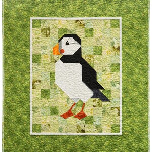 May include: A quilted wall hanging featuring a geometric puffin design. The bird is rendered in black, white, and orange, set against a patchwork background of greens and yellows. The quilt is framed with a green border.