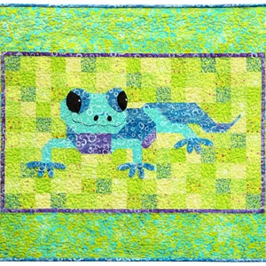 Puede incluir: Una colcha de retazos azul y verde con una rana de dibujos animados en el centro. La rana está rodeada por un borde morado y verde.