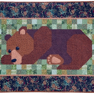 Pode incluir: Um cobertor retangular acolchoado com um desenho de urso castanho. O urso está centrado em um fundo de retalhos de quadrados verdes e azuis. A borda é um tecido azul escuro com um padrão floral.