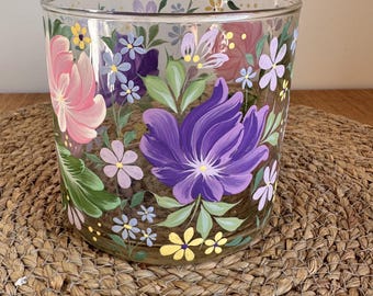 May include: A clear glass jar with hand-painted floral designs. The flowers are in shades of pink, purple, and yellow, with green leaves. The jar is sitting on a woven mat.