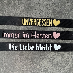 Könnte beinhalten: Drei schwarze Satinband-Lesezeichen mit weißer und goldener Beschriftung. Das obere Lesezeichen lautet "UNVERGESSEN" mit einem goldenen Herzen. Das mittlere Lesezeichen lautet "Für immer im Herzen" mit einem rosa Herzen. Das untere Lesezeichen lautet "Die Liebe bleibt" mit einem weißen Herzen.
