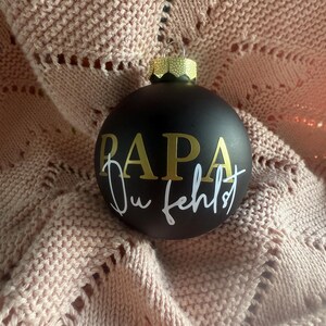 Könnte beinhalten: Eine schwarze Weihnachtskugel mit goldfarbenem Schriftzug "PAPA" und "Du fehlst" in weißer Schrift. Die Kugel hat eine goldene Kappe und liegt auf einer rosa Strickdecke. Eine festliche Weihnachtsdekoration.